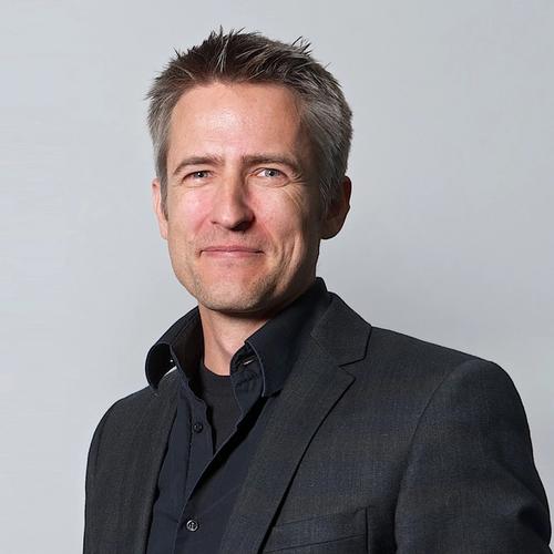  A man with graying spiky hair and blue eyes, wearing a black blazer over a black collared shirt, smiles slightly while posing for a headshot against a plain gray background.